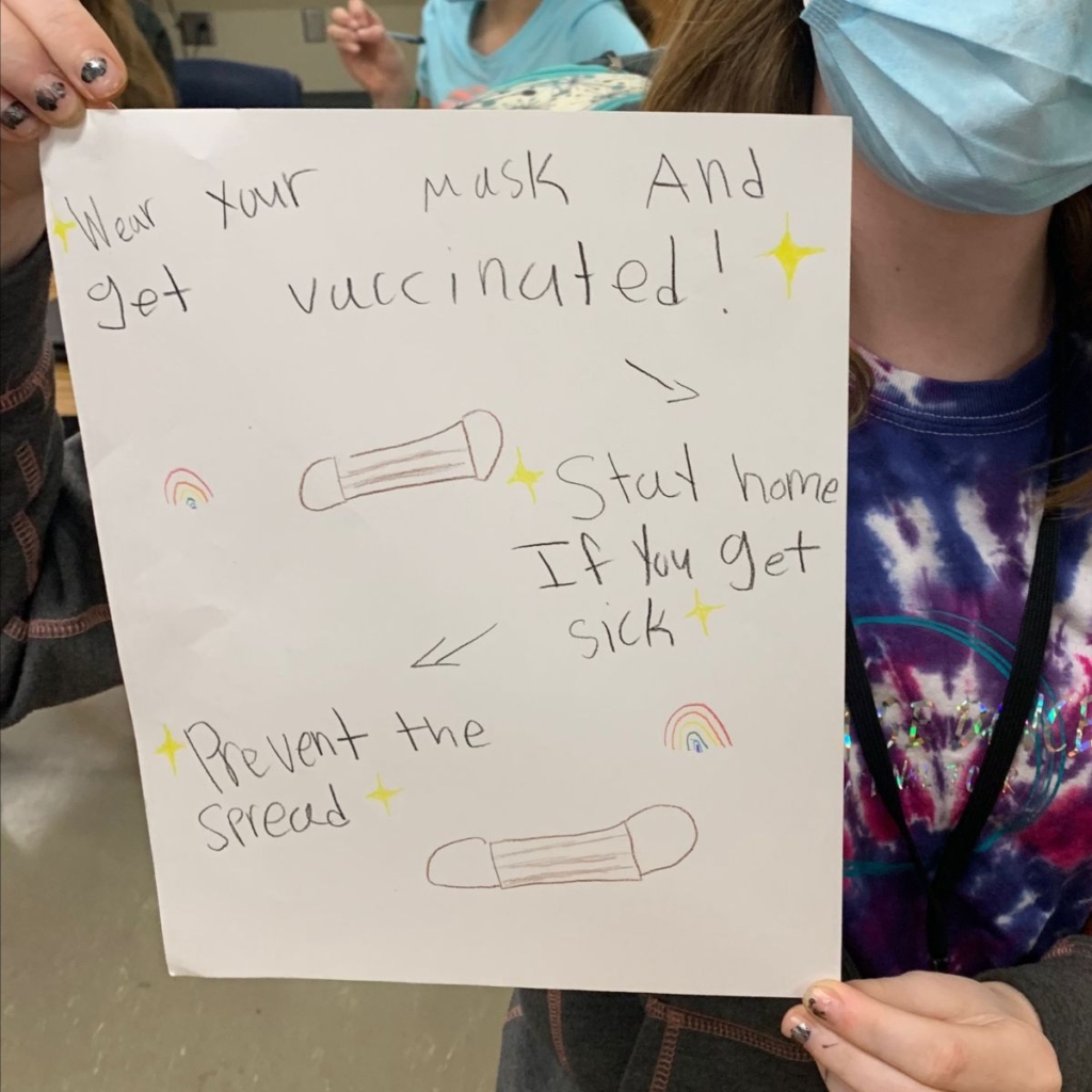 A pro-vaccination poster held up by a youth wearing a mask. The poster reads "Wear your mask and get vaccinated. Stay home if you get sick. Prevent the spread."