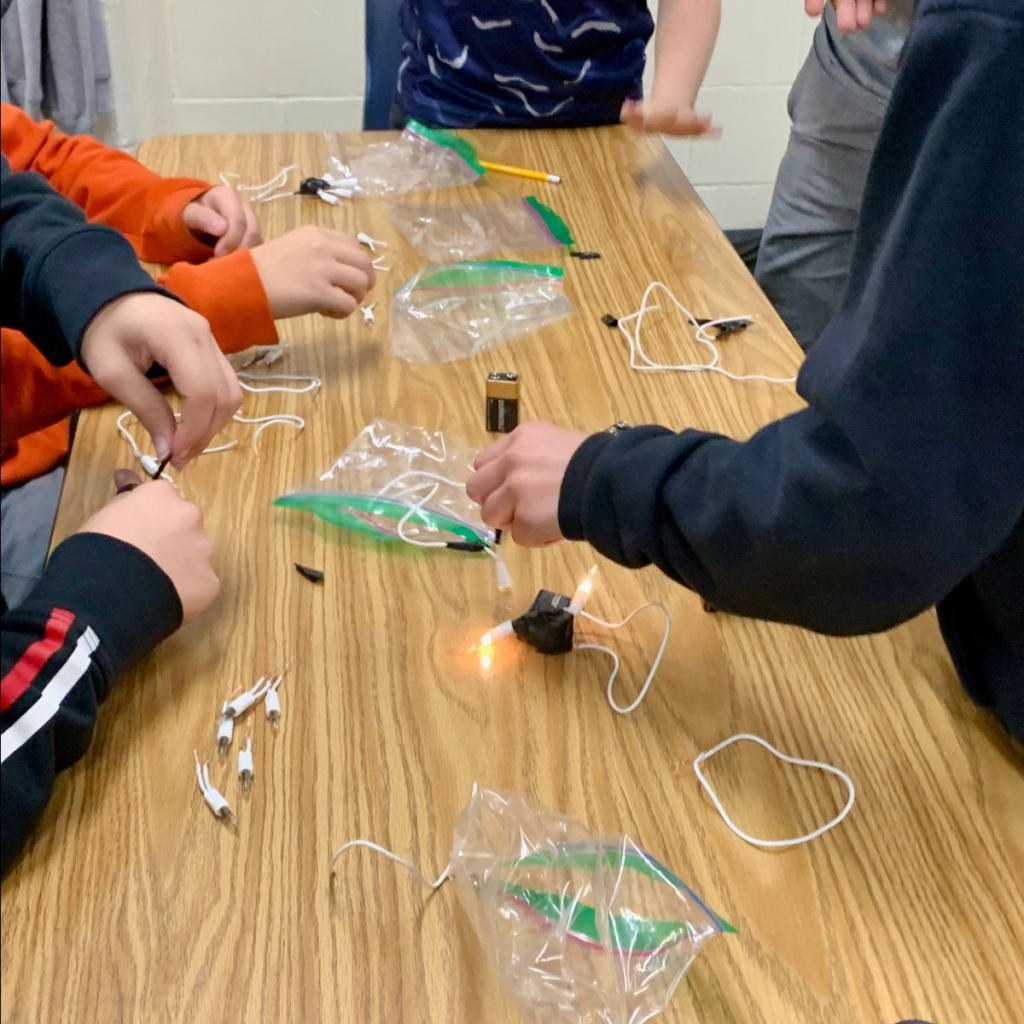 Hands of three youth with light skin building simple circuits with incandescent lights, electrical tape, and 9-volt batteries