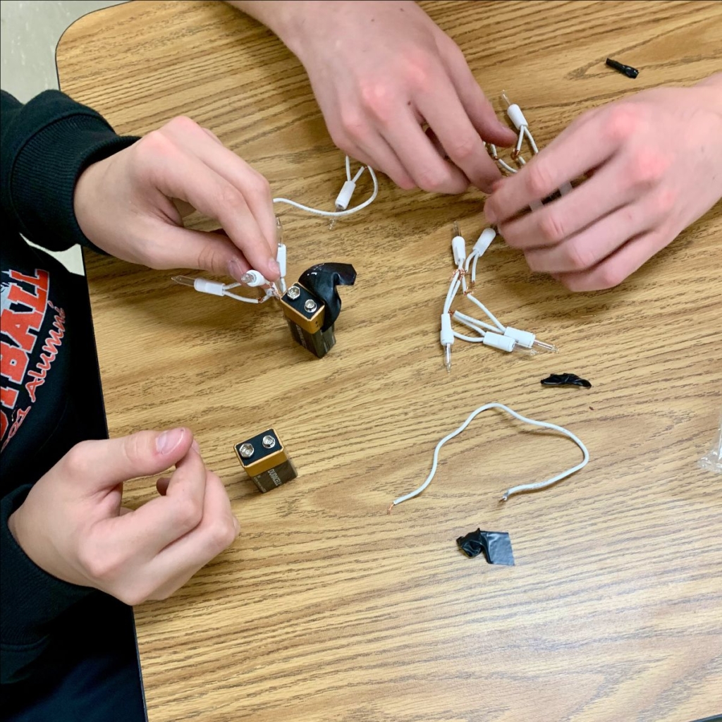 Two sets of light-skinned hands building simple circuits with incandescent lights, electrical tape, and 9-volt batteries