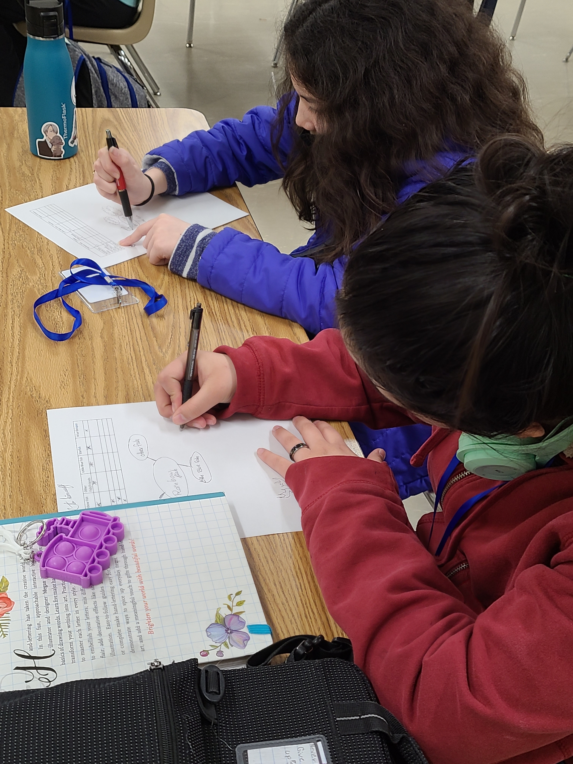 Two middle-school youth, one wearing purple and one wearing red, draw network maps on pieces of white paper.