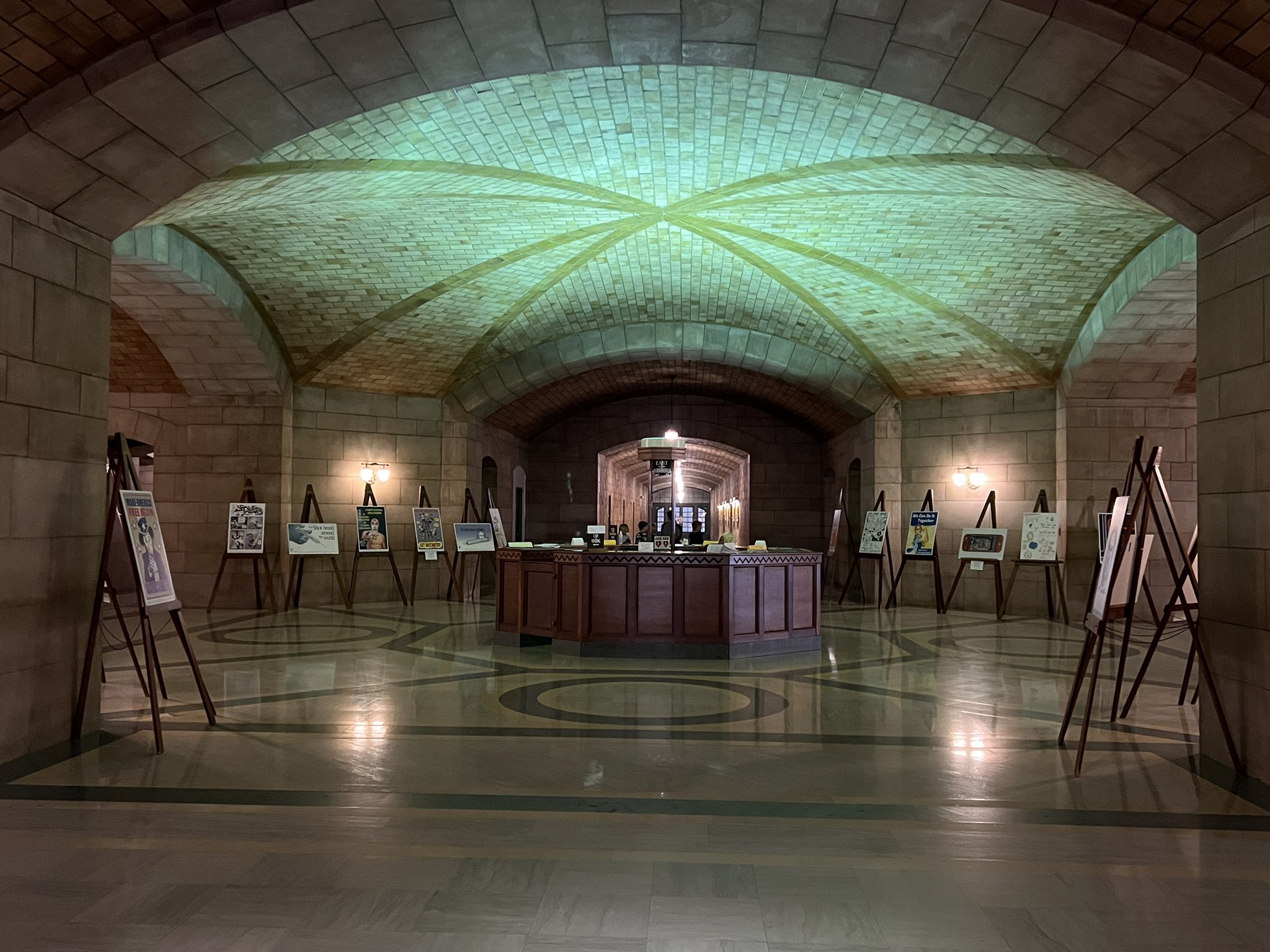 Light reflects across the ceiling of the Nebraska State Capitol first floor rotunda, creating a green glow. Posters are displayed on wooden easels around the marble interior.