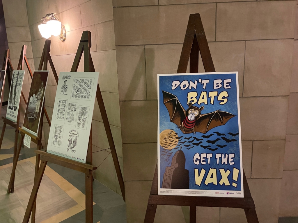 Several foam core poster boards displayed on easels against the marble background of the Nebraska State Capitol first floor rotunda. The focal poster depicts a close-up of a bat wearing a striped shirt and face covering. In the background are silhouettes of a large group of bats flying above the Capitol building against a yellow, full moon.  Poster text reads, "DON'T BE BATS / GET THE VAX!"