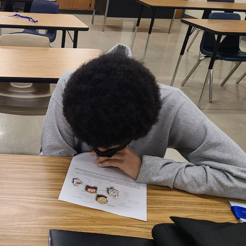 A child with brown skin wearing a grey shirt peering over a network image on a desk. The top of the youth's head is tilted toward the camera, so that their afro obscures their face.