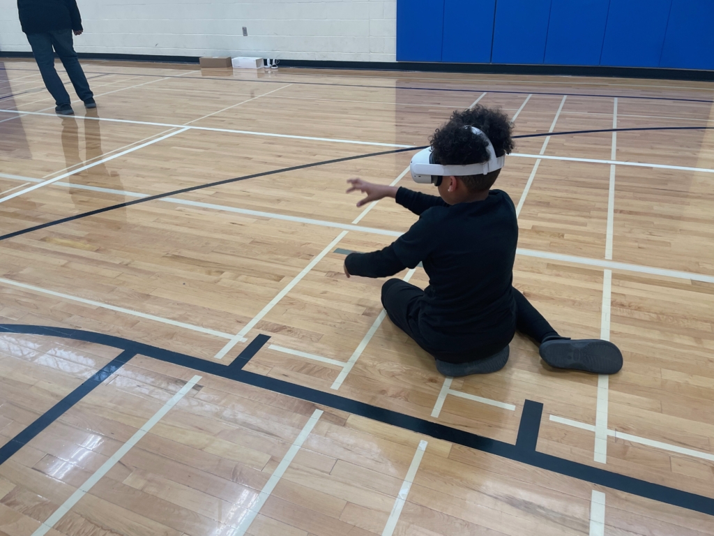 A young person with brown skin and black, curly hair sits on the floor of a middle school gym. They are wearing all-black clothes and a white VR headset, and they reach out in front of them as if to dig into the ground.