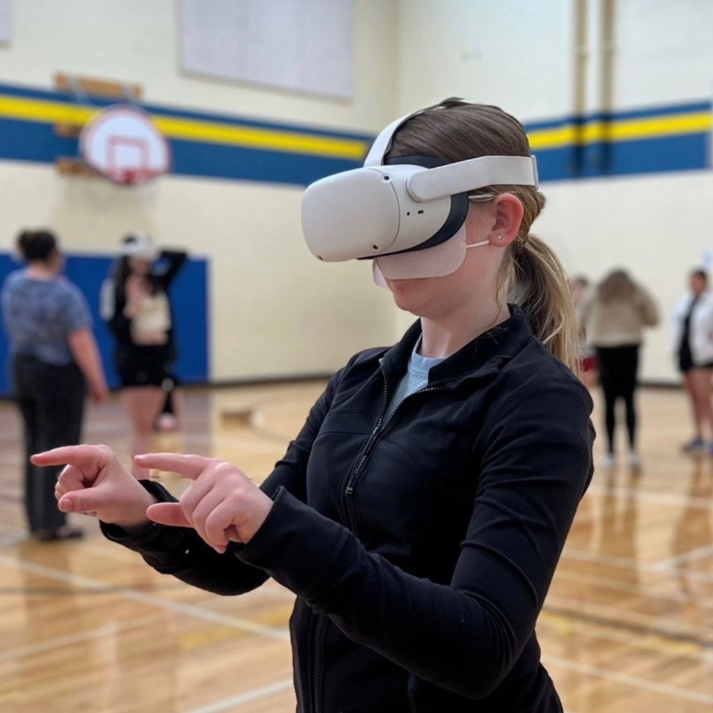 A young white person with blonde hair in a ponytail wearing a long sleeve black zipper-front shirt points forward with both fingers as they playtest a VR experience. They are wearing a white VR headset and standing in a middle school gym.