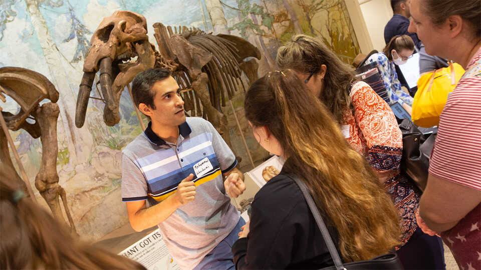 A scientist with brown skin and short, dark hair wearing a blue striped polo stands in front of a mammoth skeleton museum exhibit facing the camera. The scientist is in conversation with three teachers, who are turned away from the camera toward the discussion.